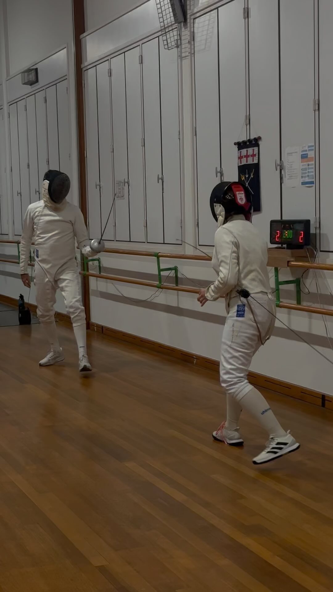 Twee schermers in vol tenue tegenover elkaar tijdens een partij, met het scorebord op de achtergrond.