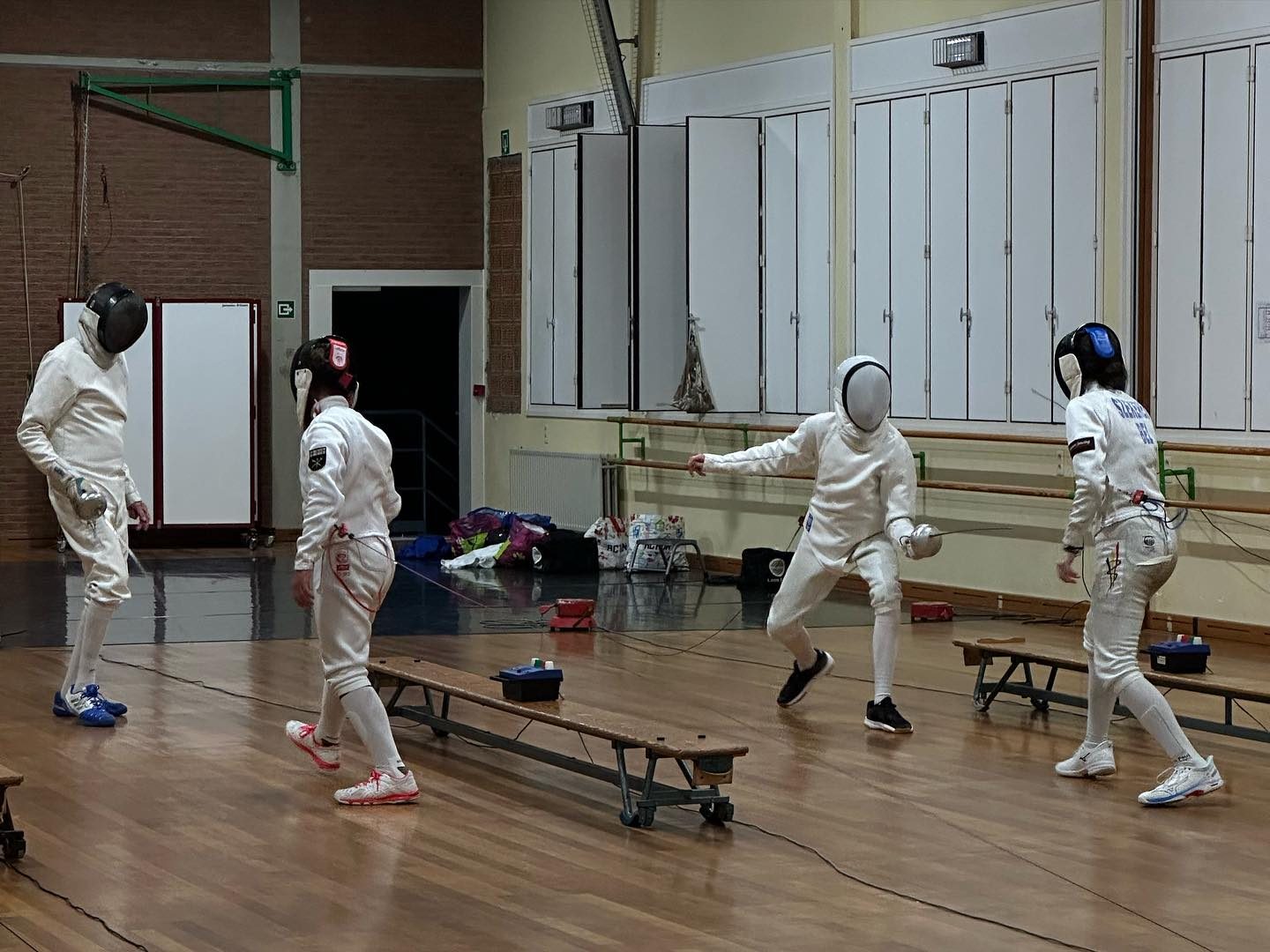 Vier schermers met masker en vest tijdens een oefening in de zaal, één van hen in uitval.