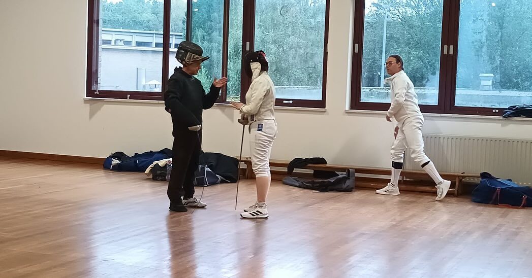 De schermleraar begeleidt twee schermers tijdens een oefening langs de ramen van de zaal.