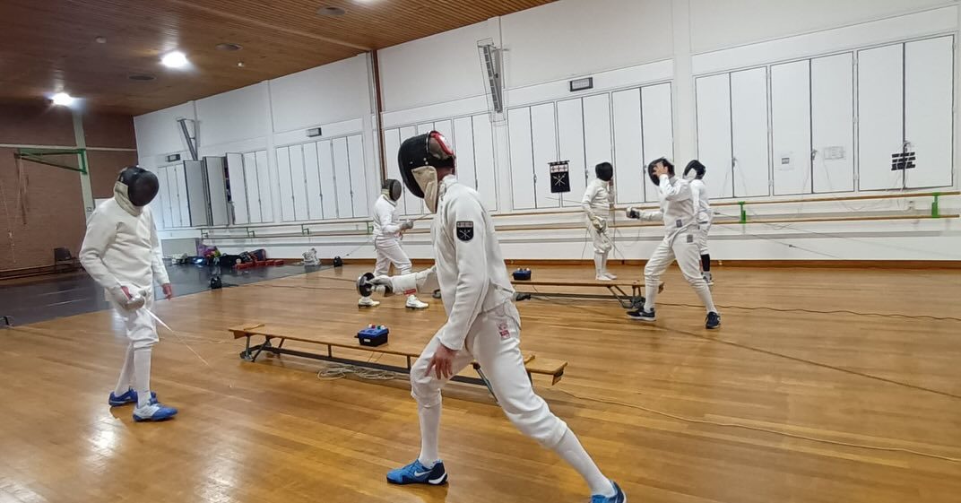 Schermers trainen verspreid over de pistes in de sporthal, gezien over de volle breedte van de zaal.